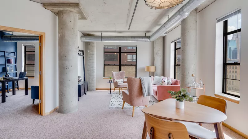 Open-plan industrial loft with pink carpet. Dining table in foreground, living area with pink sofa and chairs, office visible.