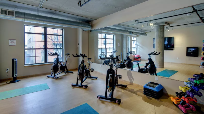 Fitness studio with stationary bikes, yoga mats, a wall mirror, and dumbbells, featuring an industrial ceiling and large windows.