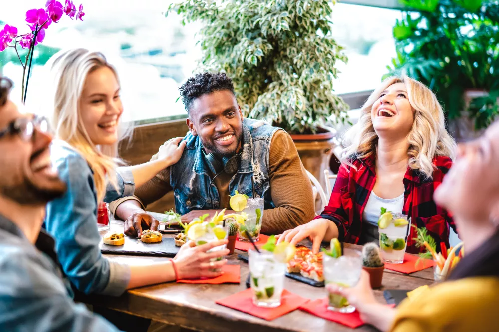 A diverse group of five friends laughing and enjoying drinks and food together at an outdoor restaurant.