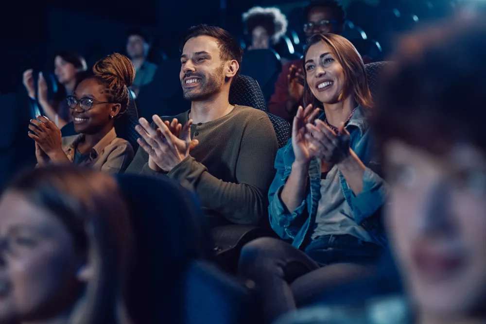 Audience members in a dimly lit movie theater smile and clap enthusiastically.